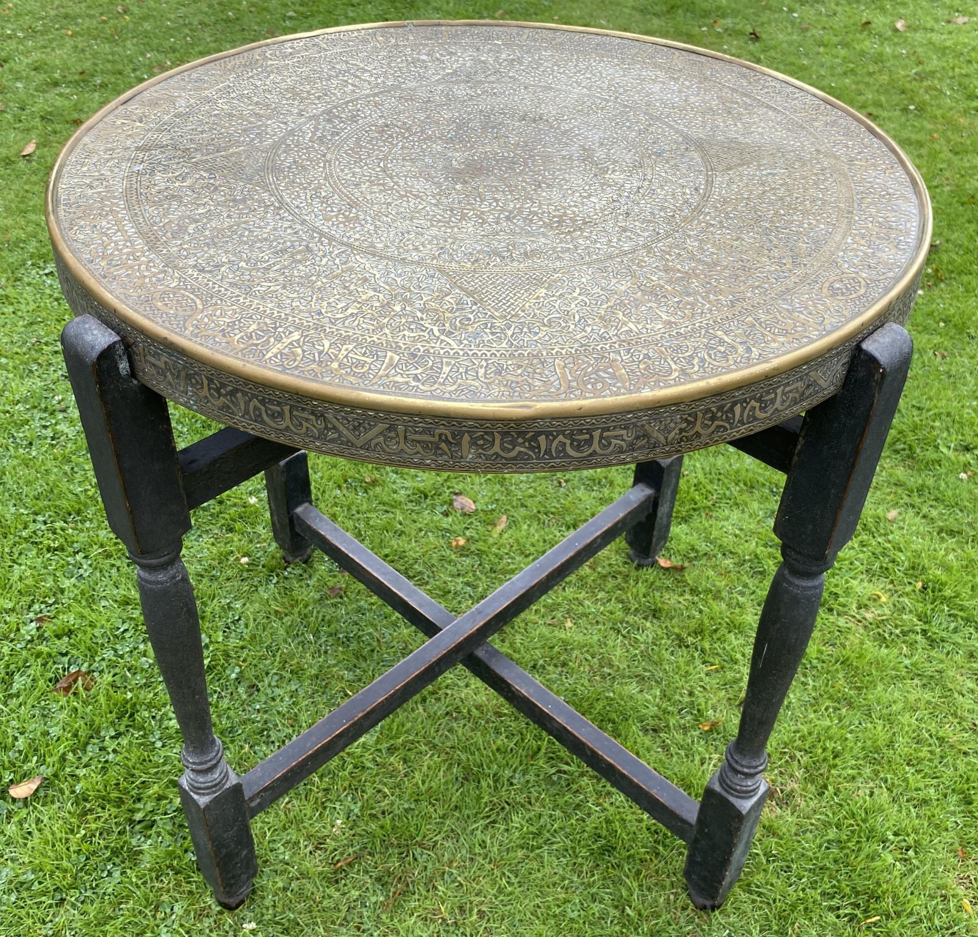 A MIDDLE EASTERN BRASS CIRCULAR TOPPED TABLE ON FOLDING WOODEN BASE, DIAMETER 59CM