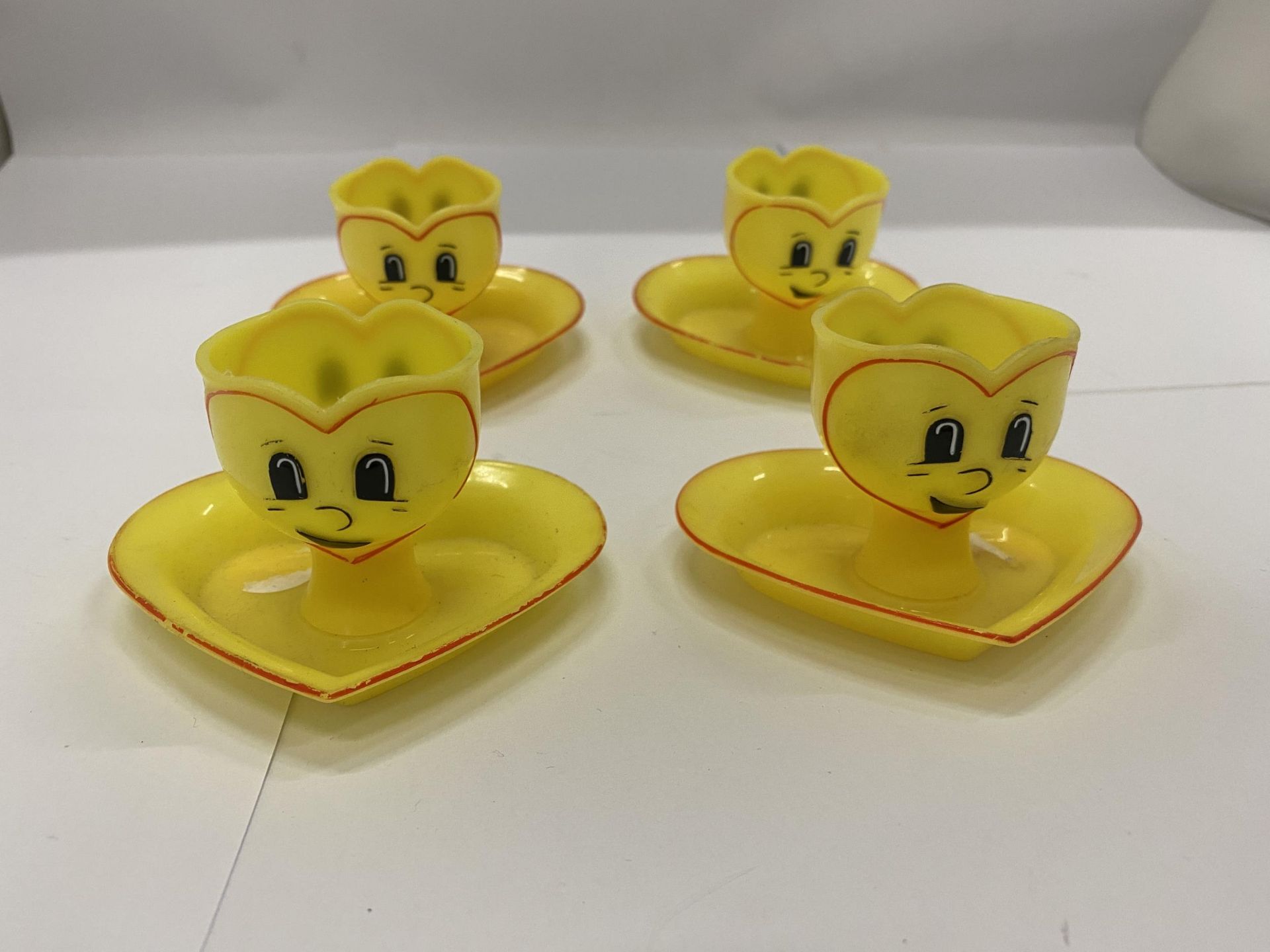 FOUR YELLOW PLASTIC EGGCUPS WITH HEART SHAPED SAUCERS AND FACES ON THE FRONT AND BACK