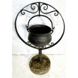 A cast iron cauldron in a wrought iron hanging stand with concrete base.