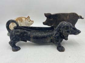 A cast iron boot scraper in the form of a dachshund along with two door stops in the form of pigs