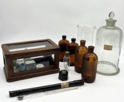 A collection of chemists bottles and bell jars along with a small wooden medical cabinet