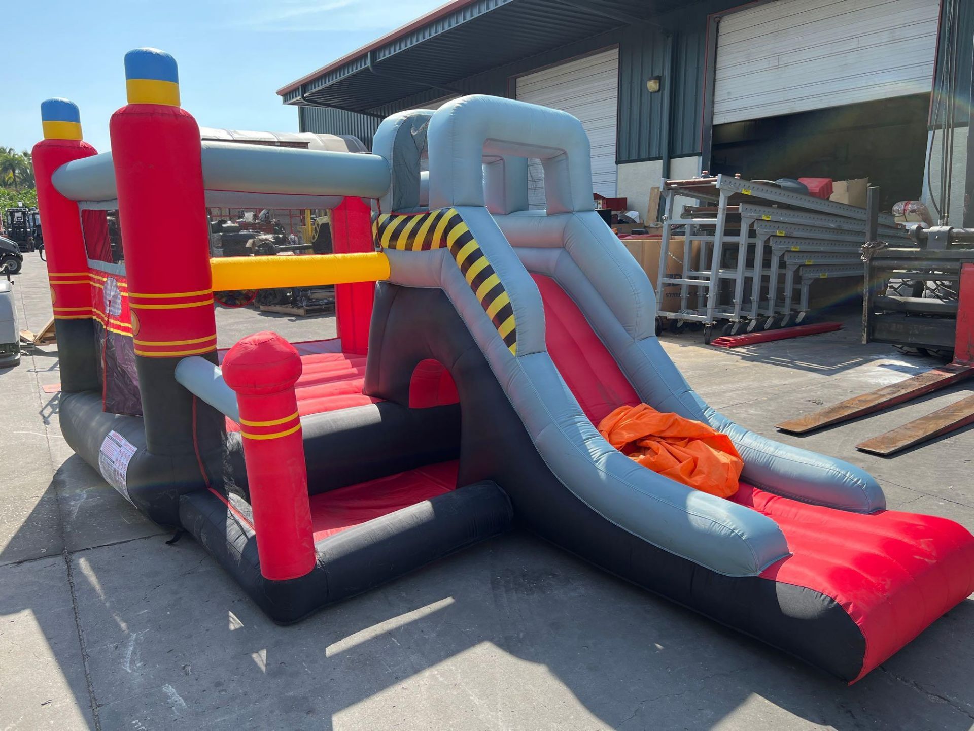 UNUSED BLOW-UP BOUNCY FIRE STATION PLAY YARD WITH SLIDE, VELCRO DARTS, BALLS, STAKES, & BLOWER