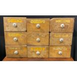 A set of nine pine apothecary drawers, porcelain handles, paper labels, 23cm wide x 14.5cm high x