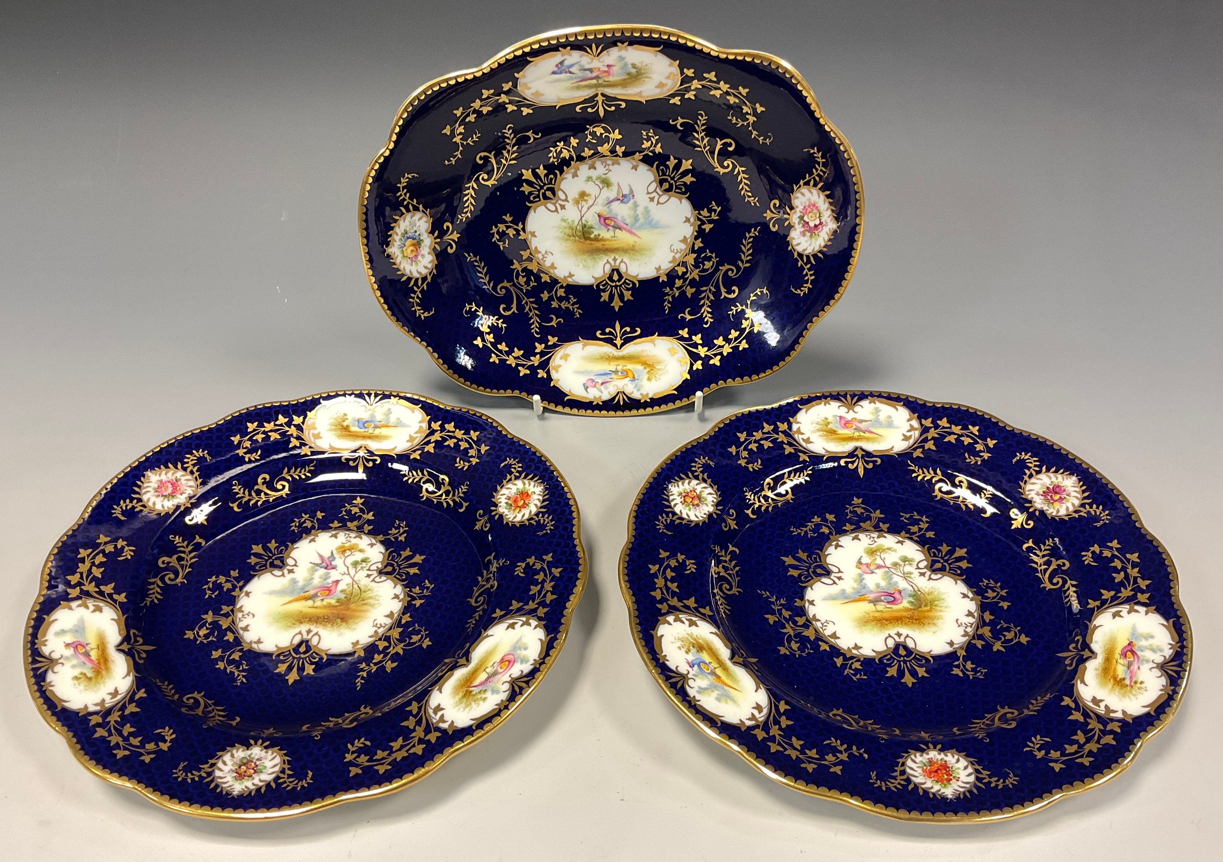 A Coalport oval dish and pair of circular plates painted with panels of birds and flowers, cobalt