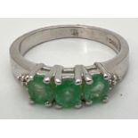 A Genuine Gem Company silver and emerald trilogy ring with diamond clusters set to each shoulder.