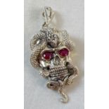 An unmarked white metal pendant in the form of a skull with a snake, set with rubies for eyes.