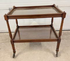 A vintage dark wood tea trolley with turned legs and glass inserts to shelves.