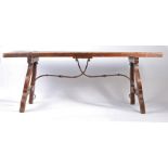 A Spanish elm refectory table, the two plank top above angled supports united by a scrolling iron