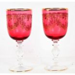 Pair of cranberry and gilt decorated wine glasses, with floral garland decoration, 18cm tall,