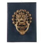 A cast brass lion mask door knocker, 18cms diameter, mounted on a wooden plaque.