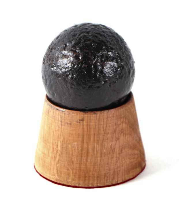 A roundshot iron cannon ball. Approximate diameter of 6.5cms (2.5ins) on a turned oak mount