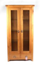 A modern oak display cabinet, the pair of glazed doors enclosing a shelved interior above a single