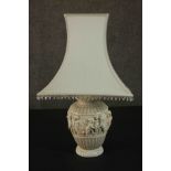 An ivory coloured resin table lamp, cast with a Bacchanalian scene, with a shade. H.67 W.40cm.