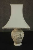 An ivory coloured resin table lamp, cast with a Bacchanalian scene, with a shade. H.67 W.40cm.