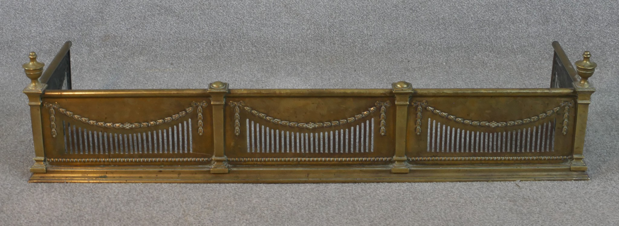 An Adam style pierced brass fender, with urn finials to the corners, decorated with swags and a