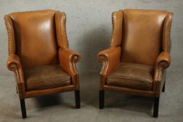 A pair of Georgian style tan leather wing back armchairs, with scrolling arms and studded detail, on