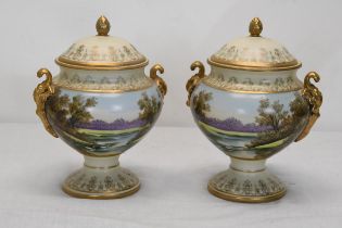 A pair of hand painted lidded twin handled ceramic urns with lake scene with swans and gilded
