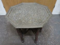 AN ORIENTAL BRASS TOPPED FOLDING TABLE, the folding base with extensive carved decoration, the
