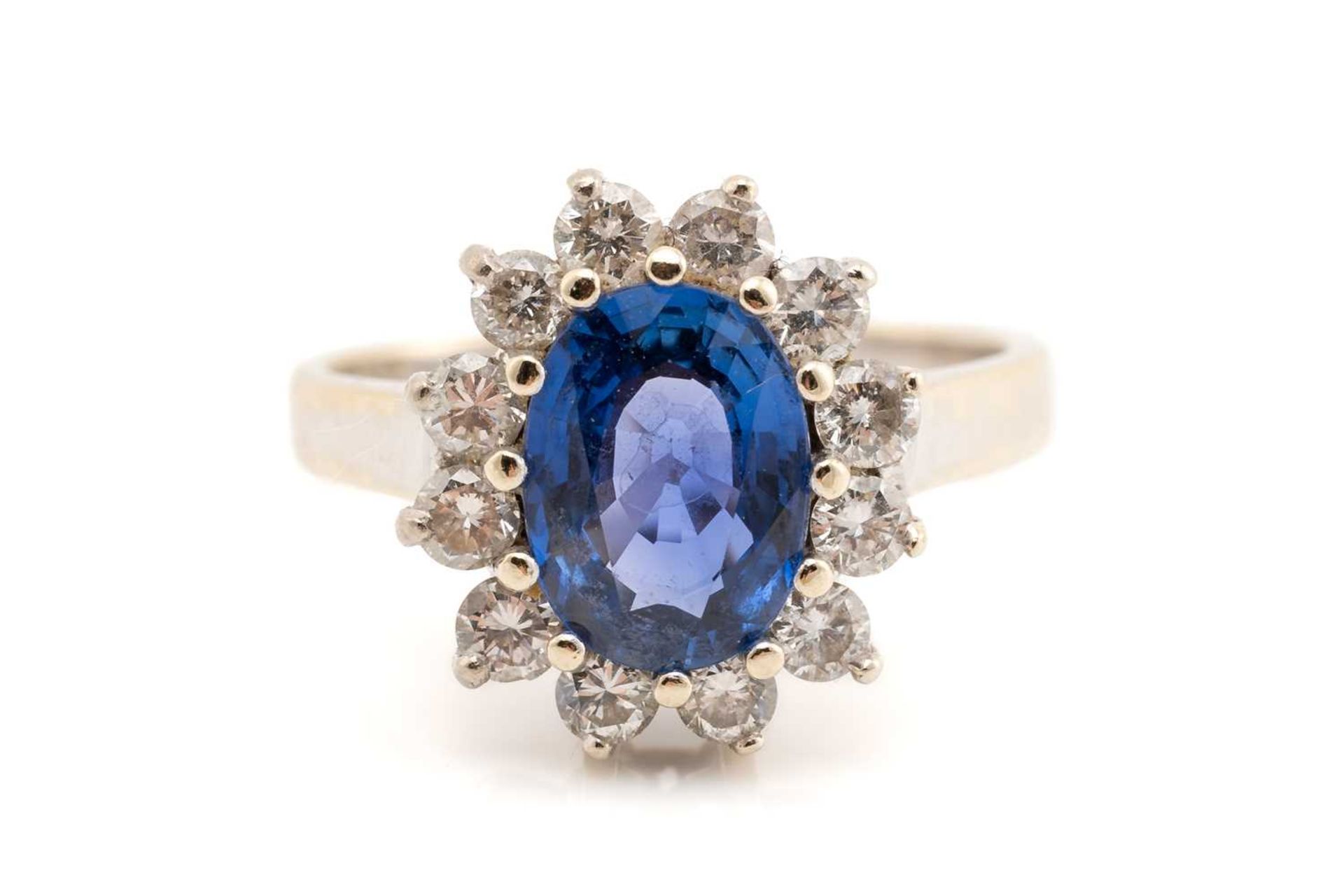 A sapphire and diamond cluster ring; the oval cut sapphire in claw mount above a border of twelve