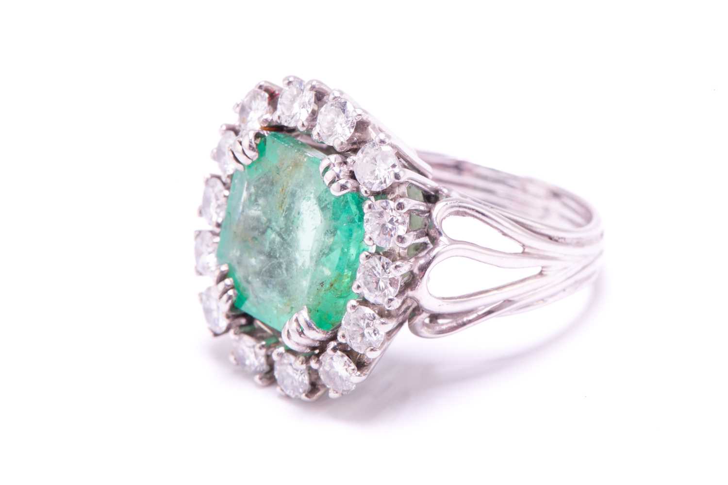 An emerald and diamond entourage ring, centred with an emerald-cut emerald of pale bright bluish- - Image 2 of 12