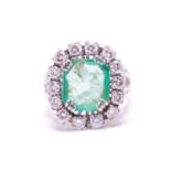 An emerald and diamond entourage ring, centred with an emerald-cut emerald of pale bright bluish-