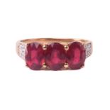 A yellow metal, diamond, and ruby ring set with three mixed oval-cut rubies, the shoulders pave