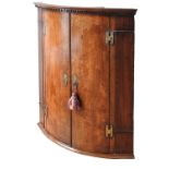 A GEORGE III OAK 19TH CENTURY BOW FRONT CORNER CUPBOARD, the two oak doors flanked by cross banded