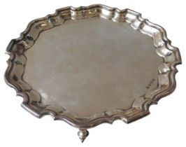 A SILVER SALVER, circular scroll edge form, raised on three scrolling feet, bears the mark HJL,