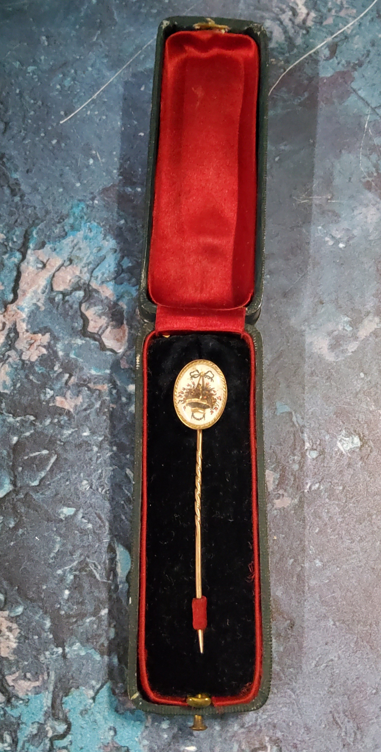 A gold coloured metal stick pin, the oval terminal set with basket of flowers, cased