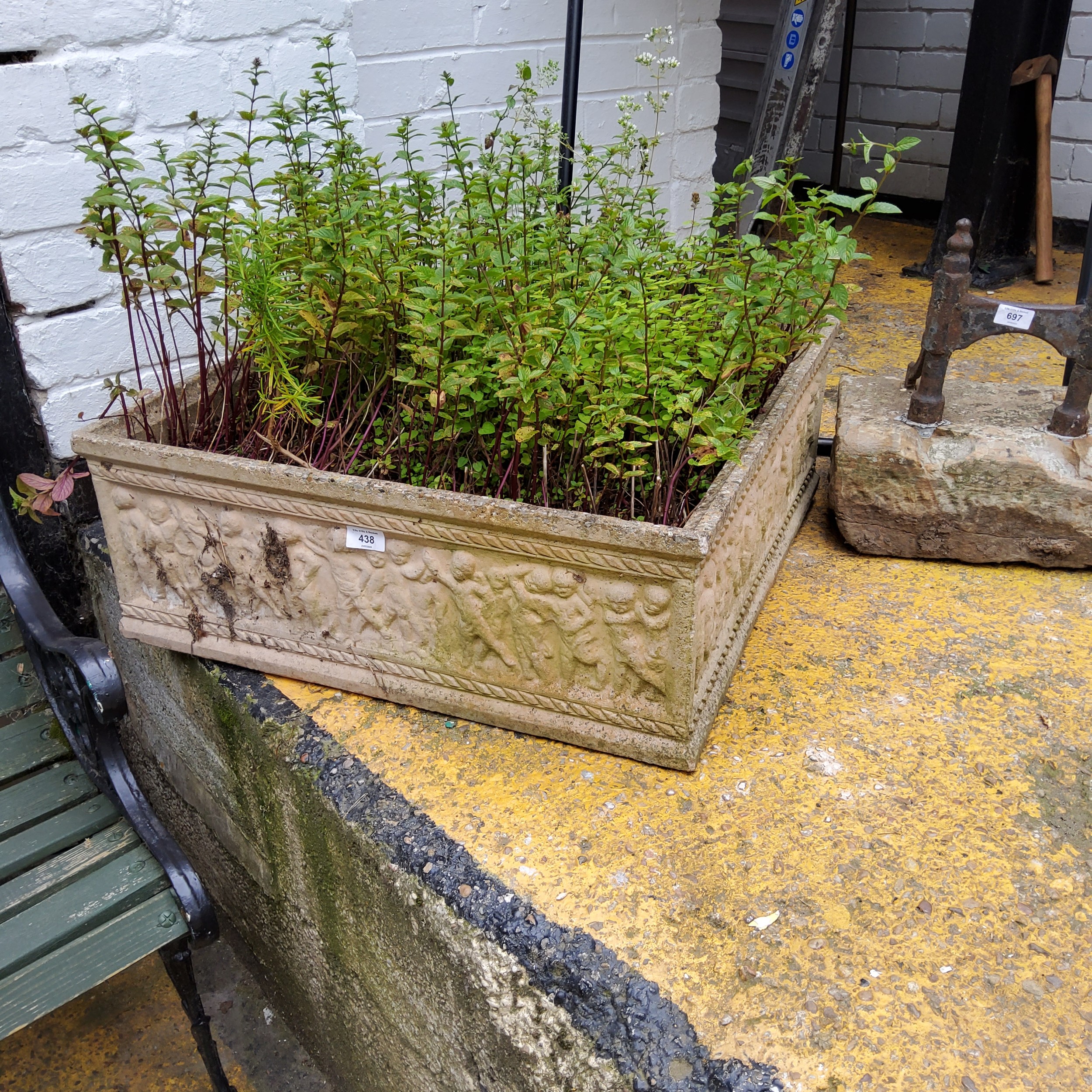 A large square reconstituted stone planter, decorated in relief with putti figures, with herbs