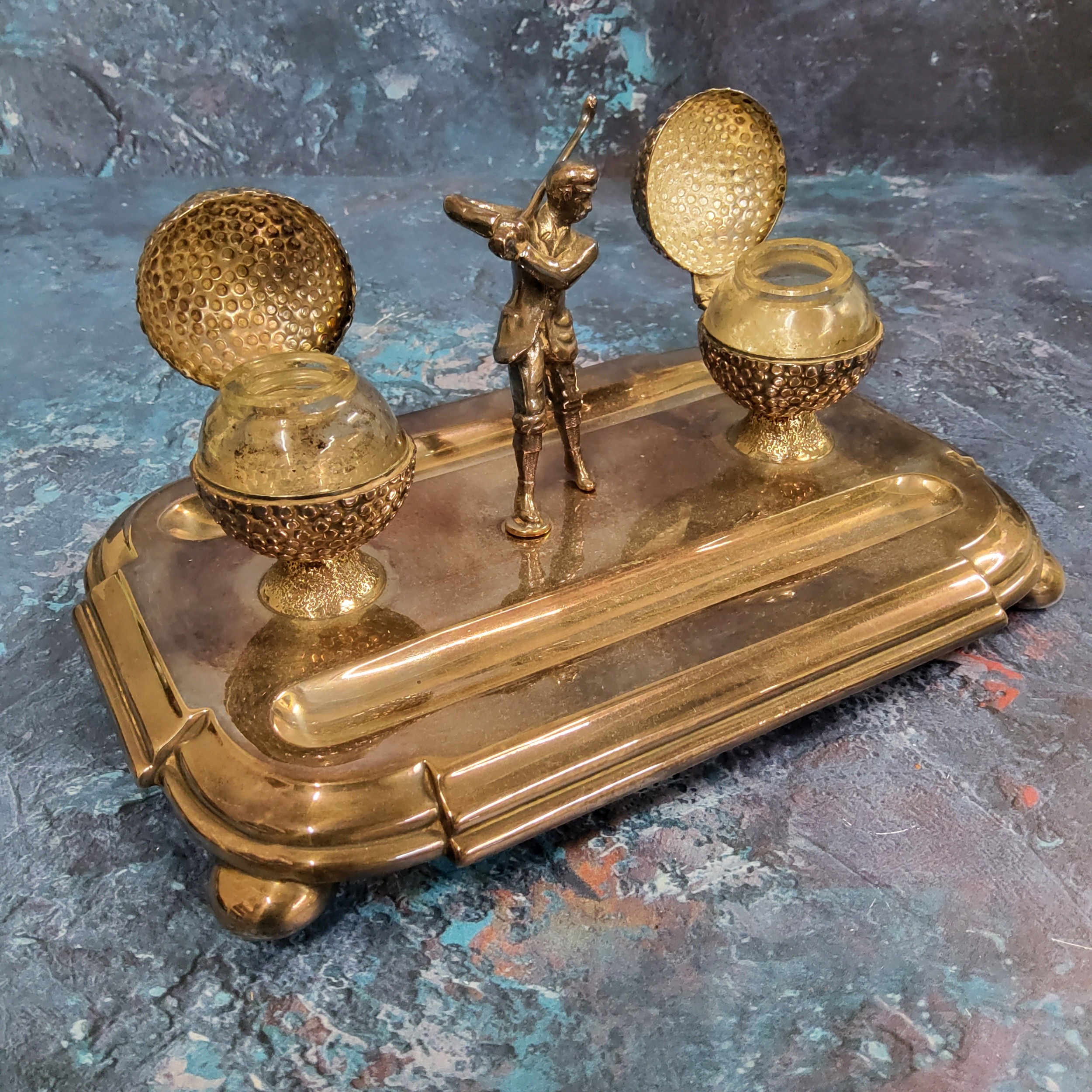 A silver plated desk standish, with golfer flanked by two golf ball wells, pen apertures, bun