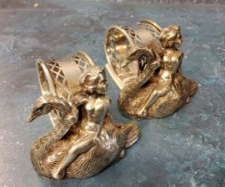 A pair of plated novelty napkin rings, cast with cherubs riding swans, 7cm wide