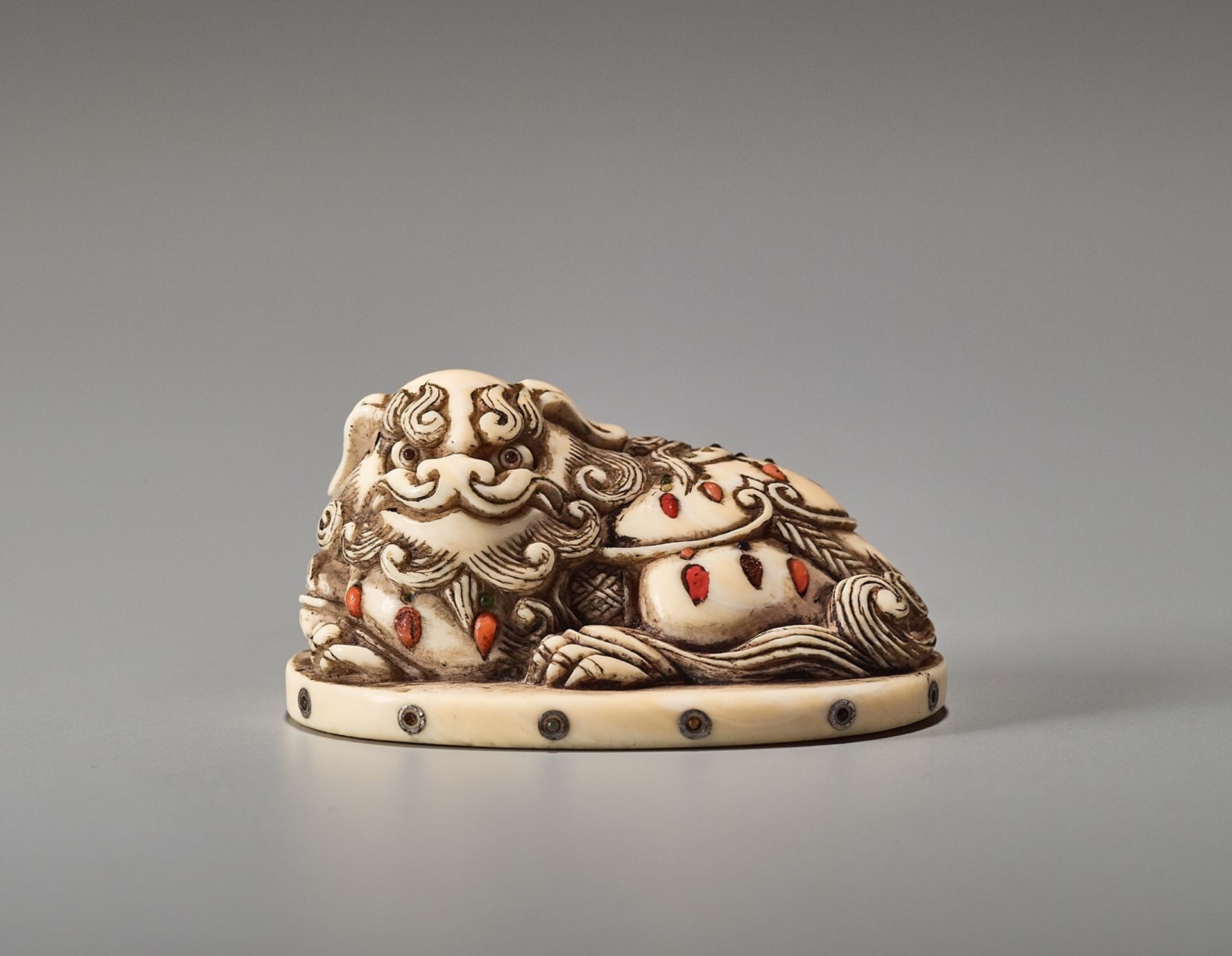 AN INLAID IVORY NETSUKE OF A RECUMBENT SHIBAYAMA SHISHI