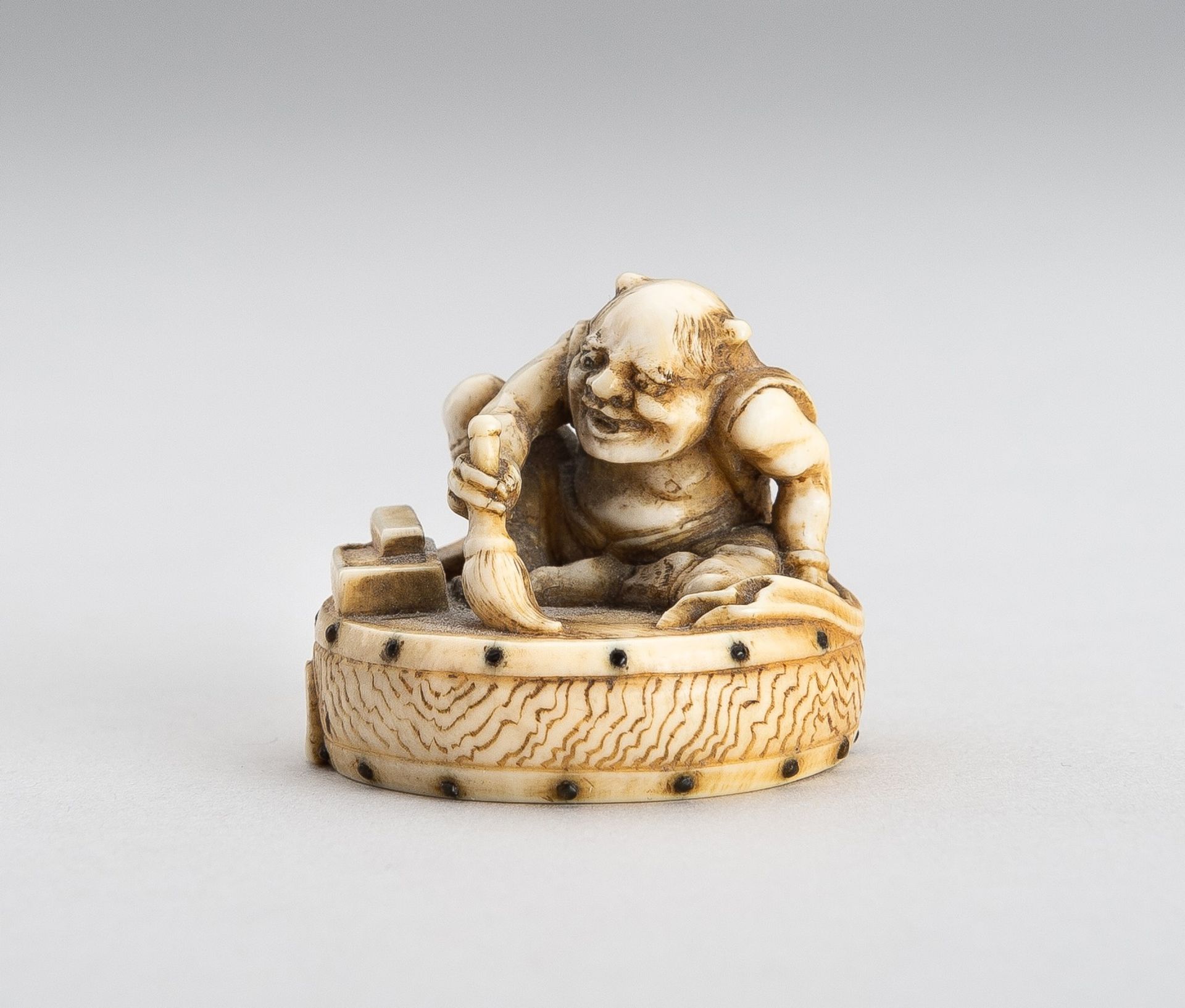 GYOKUSHU: AN IVORY NETSUKE OF AN ONI CLEANING A DRUM