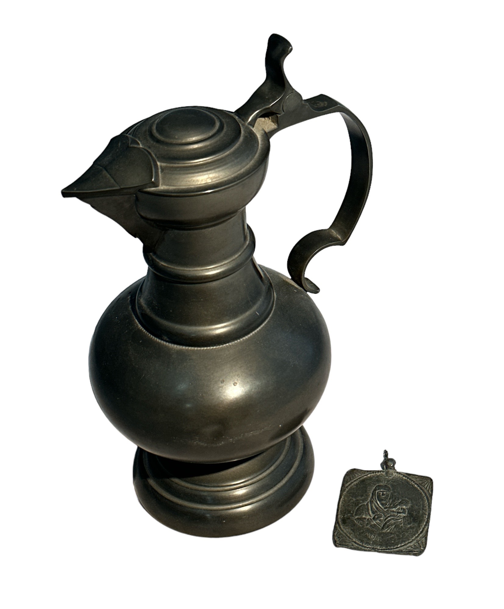 Pewter lidded jug (height 26cm) with two-sided religious token (possibly lead) 6cm x 5cm.