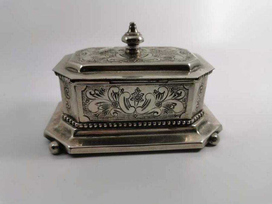 A white metal canted rectangular cigarette box with foliate scrolling design, ball feet, Arabic