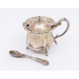 A George V silver mustard pot and cover, flared rim with foliate decoration, on three foliate and