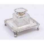 An Edwardian silver mounted cut glass ink bottle on stand, hinged cover with scrolling decoration,