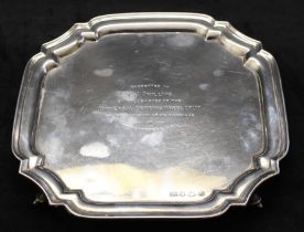 A silver shaped square salver with raised moulded border and on four scroll feet, the centre with