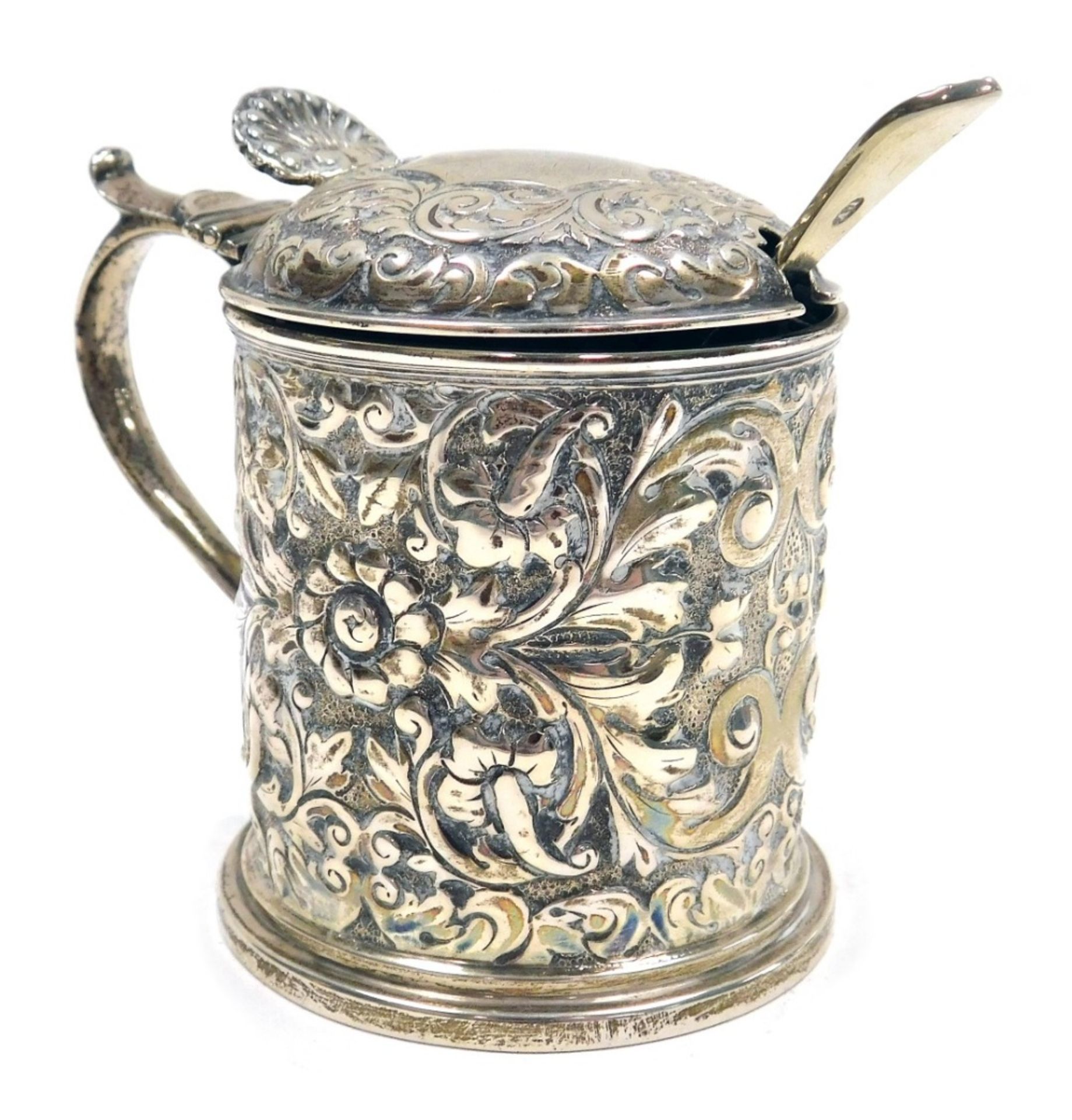 A Victorian silver mustard pot, the body and lid embossed with flowers, leaves and scrolls with a sh