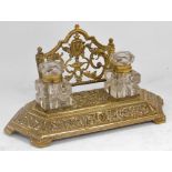 A Victorian brass desk stand, with embossed foliate scroll decoration, the top having two cut glass