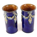 A pair of Royal Doulton cylindrical vases, each decorated with flower, leaf and grape swags, on a bl