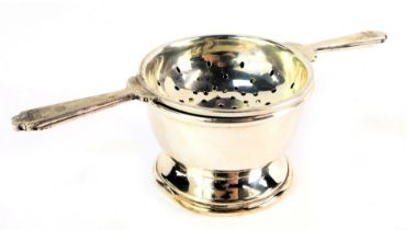 An Elizabeth II silver tea strainer and cup, the tea strained with Art Deco style handles, on a plai