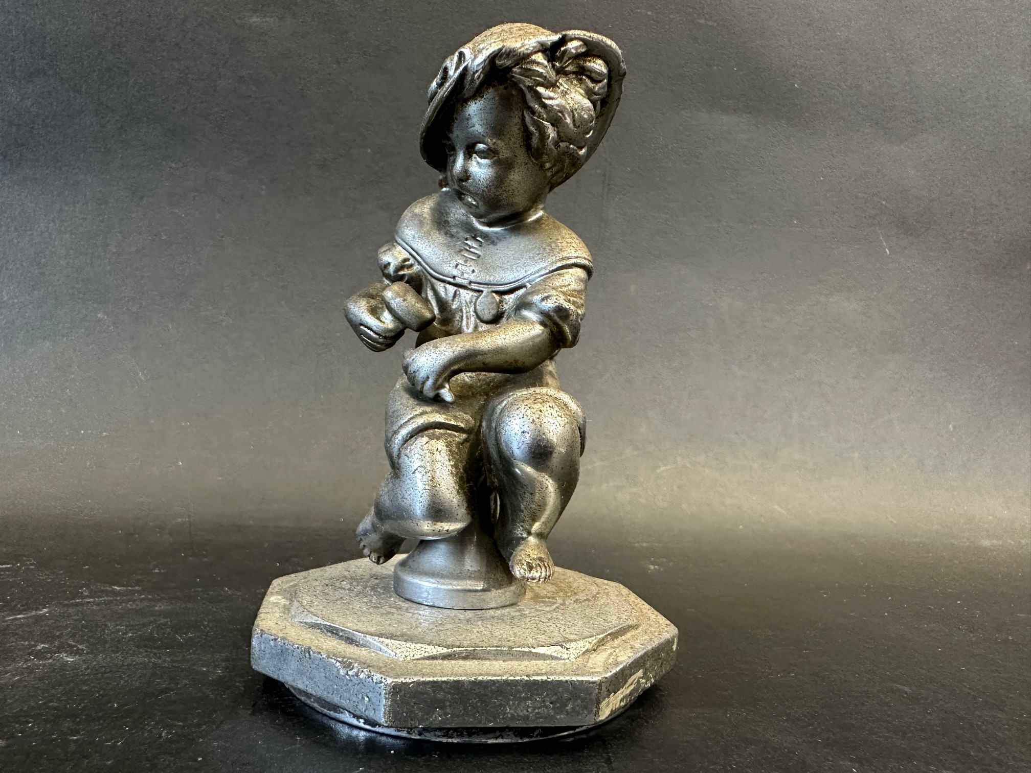 A car mascot in the form of a seated child holding a hammer and chisel, radiator cap mounted,