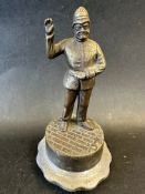 A bronze car mascot in the form of a policeman holding up his right hand and stood on a cobbled