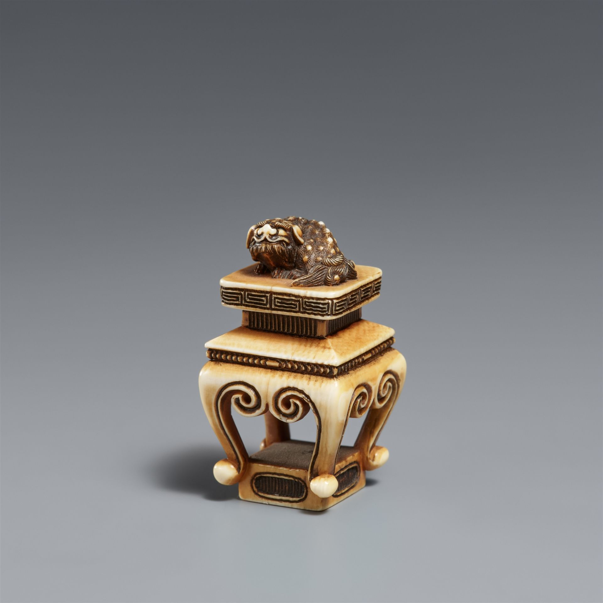 A stained ivory netsuke of a shishi on a Chinese table. Osaka. Around 1860