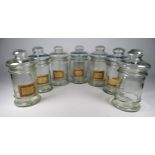 Seven clear glass apothecary jars - some with paper contents labels, with button type covers, height