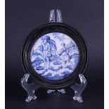 A framed earthenware tile, round with a decoration of two figures in a landscape.