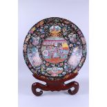 A very large famille noir dish decorated with various figures, in a wooden stand. China, 20th centur