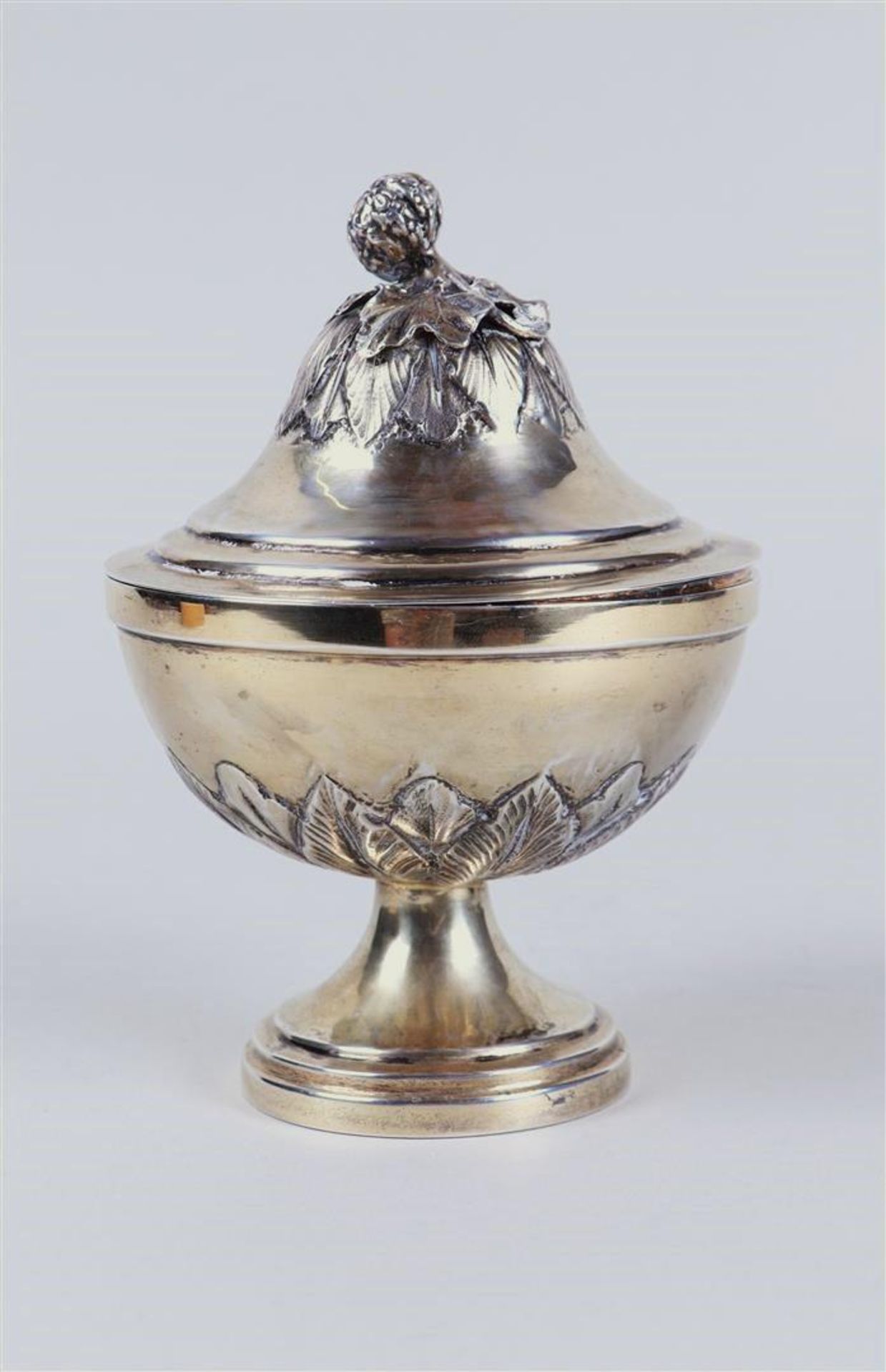 A silver lidded coupe on a round base with a constricted base surrounded by palm leaves, the lid wit